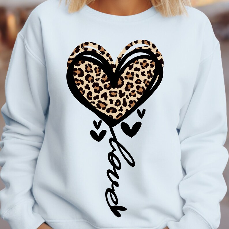 Peut inclure: Un sweat-shirt bleu clair avec un motif de c&oelig;ur. Le c&oelig;ur est bord&eacute; de noir et pr&eacute;sente un motif l&eacute;opard &agrave; l'int&eacute;rieur. En dessous, le mot "Love" est &eacute;crit en cursive, avec de petits c&oelig;urs noirs.