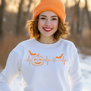 May include: White sweatshirt with an orange pumpkin and bat graphic. The graphic features a heartbeat line with a pumpkin in the centre and bats on either side. The text "Boo" is written above the graphic.