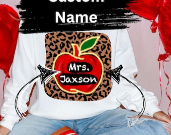 Pode incluir: Camisola branca com um gráfico de maçã vermelha sobre um fundo de estampa de leopardo. A maçã tem o texto "Mrs. Jaxson" escrito nela.