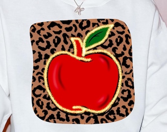 Pode incluir: Camisola branca com um gráfico de maçã vermelha sobre um fundo de estampa de leopardo marrom. A maçã tem um contorno dourado e uma haste e uma folha verdes.