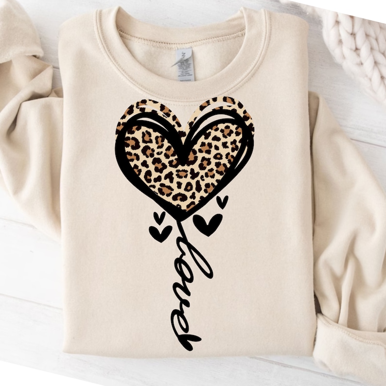 Peut inclure: Un sweat-shirt beige avec un grand motif de c&oelig;ur. Le c&oelig;ur est soulign&eacute; en noir et rempli d'un motif l&eacute;opard. Le mot "Love" est &eacute;crit en cursive en dessous du c&oelig;ur. De petits c&oelig;urs noirs sont &eacute;galement pr&eacute;sents.