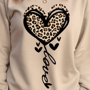 May include: A beige sweatshirt featuring a large black outlined heart with a leopard print design. Below the heart, the word "Love" is written in a cursive font, with small black hearts accenting the design. The sweatshirt has a crew neck.