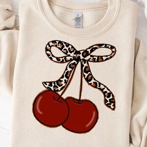 May include: A cream-colored sweatshirt with a graphic of two red cherries with a leopard print bow on top.