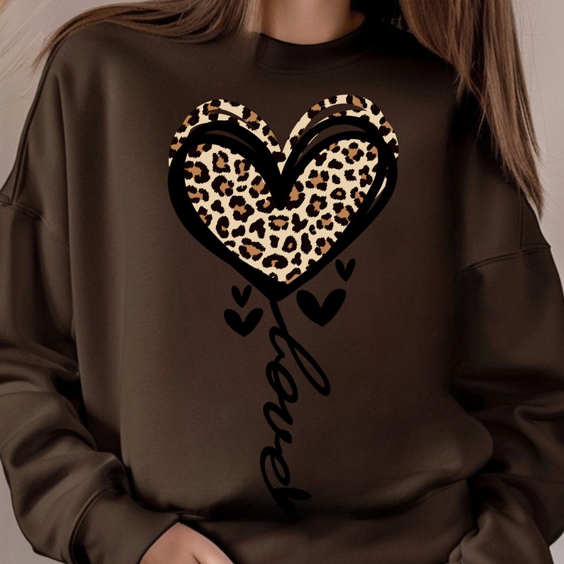 Peut inclure: Un sweat-shirt marron avec un motif de c&oelig;ur. Le c&oelig;ur pr&eacute;sente un imprim&eacute; l&eacute;opard et est soulign&eacute; en noir. Le mot "Love" est &eacute;crit en cursive en dessous du c&oelig;ur, avec de petits c&oelig;urs.