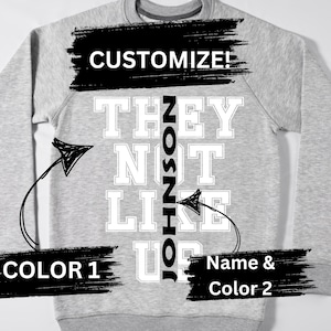 Puede incluir: Sudadera gris con el texto "CUSTOMIZE!" en la parte superior. El texto "THEY NOT LIKE US" está impreso en letras blancas con una línea vertical negra en el medio. El texto "ZOSZHIO" está escrito verticalmente en letras negras sobre la línea negra. Las flechas apuntan al texto.