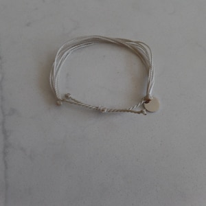 May include: A white string bracelet with a silver charm. The bracelet is made of multiple strands of string and has a simple design.