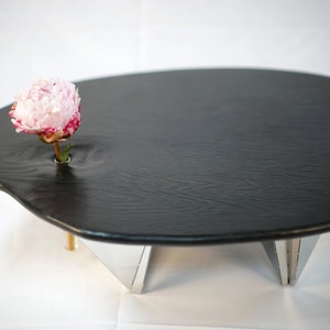 Op de afbeelding: Een zwarte, onregelmatig gevormde tafel met een enkele roze bloem in een gat in het midden van het tafelblad. De tafel wordt ondersteund door drie zilveren metalen poten.