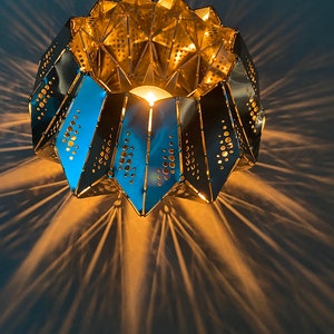 Puede incluir: Un portavelas de metal dorado con un diseño geométrico y una pequeña vela en su interior. El portavelas proyecta un patrón de luz en forma de estrella sobre la superficie de abajo.