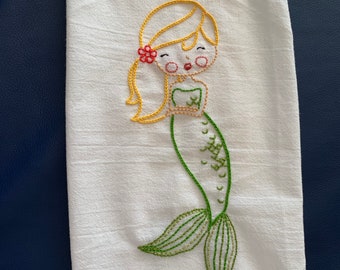 May include: A white kitchen towel with an embroidered mermaid design. The mermaid has a green tail, yellow hair, and a red flower in her hair.