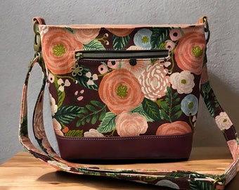 May include: A floral patterned crossbody bag with a burgundy leather bottom and a gold zipper. The bag has a long adjustable strap.