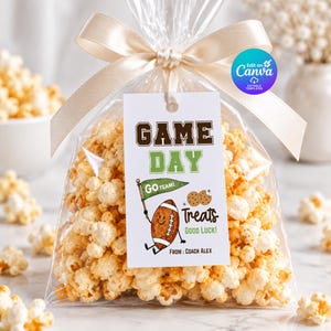 Puede incluir: Una bolsa de plástico transparente llena de palomitas de maíz, atada con una cinta de satén beige. Una etiqueta rectangular blanca muestra el texto GAME DAY, GO TEAM, Treats, GOOD LUCK y FROM: COACH ALEX, con dibujos de un balón y galletas.