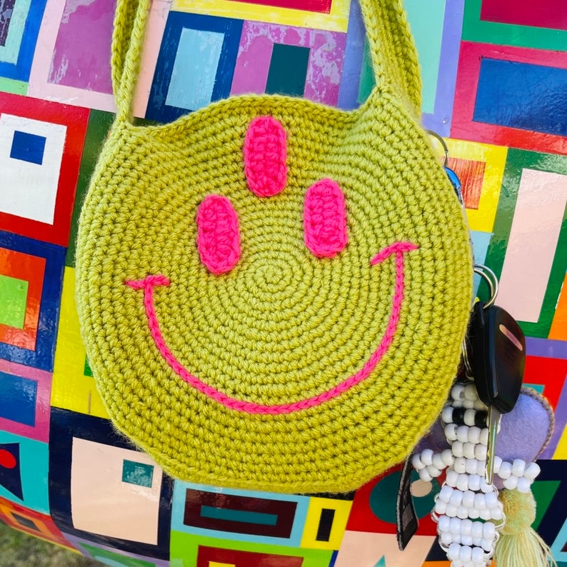May include: A green crocheted bag with a pink smiley face design. The bag has a long strap and is hanging on a colorful background.