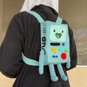 Puede incluir: Una mochila de ganchillo azul claro con una cara de dibujos animados y piernas. La mochila tiene forma cuadrada con una cara verde, una estrella amarilla, un círculo rojo y formas azules y naranjas. Las correas son de color azul claro y las letras "BM" están en el lateral.