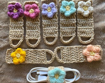 Puede incluir: Una colección de organizadores de cables y enrolladores de cordón hechos a mano con ganchillo. Los organizadores son de color beige con acentos florales coloridos en morado, rosa, azul, verde, amarillo y melocotón. El enrollador de cable está alrededor de un cable blanco.