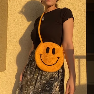 May include: A person wearing a black shirt and a black and white skirt with a celestial print is carrying a yellow crocheted smiley face bag.