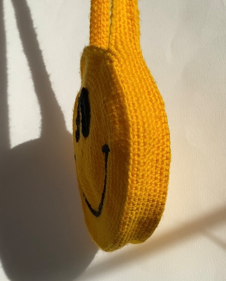 May include: A yellow crocheted smiley face bag with a black stitched smile and eyes. The bag has a yellow crocheted strap.