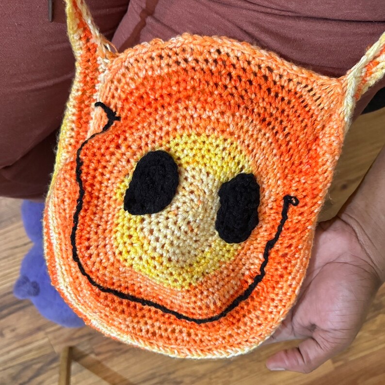 May include: Orange and yellow crocheted smiley face bag with black yarn for the eyes and mouth.