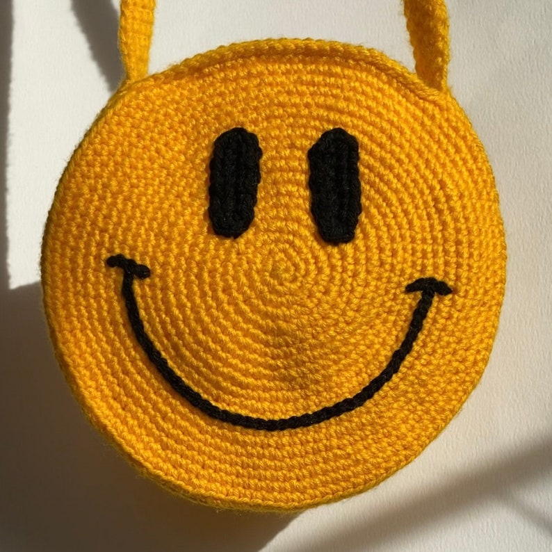 May include: A yellow crocheted bag shaped like a smiley face with a black outline. The bag has a black strap.