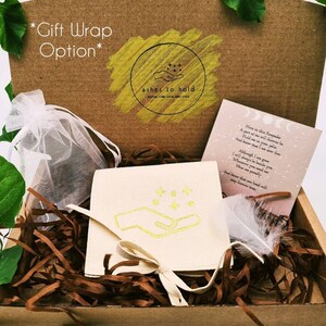 Pode incluir: Uma caixa de presente com uma bolsa creme com um design de m&atilde;o e estrelas douradas, um cart&atilde;o com um poema e uma pena branca. A caixa tamb&eacute;m cont&eacute;m enchimento de papel castanho e um saco branco transparente. O texto "ashes to hold" &eacute; vis&iacute;vel.