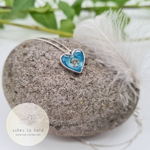 Pode incluir: Um pingente prateado em forma de cora&ccedil;&atilde;o com um centro de resina azul, contendo uma pequena quantidade de material de cor clara. O pingente pende de uma delicada corrente de prata. O colar repousa sobre uma pedra cinza, com uma pena branca por perto.