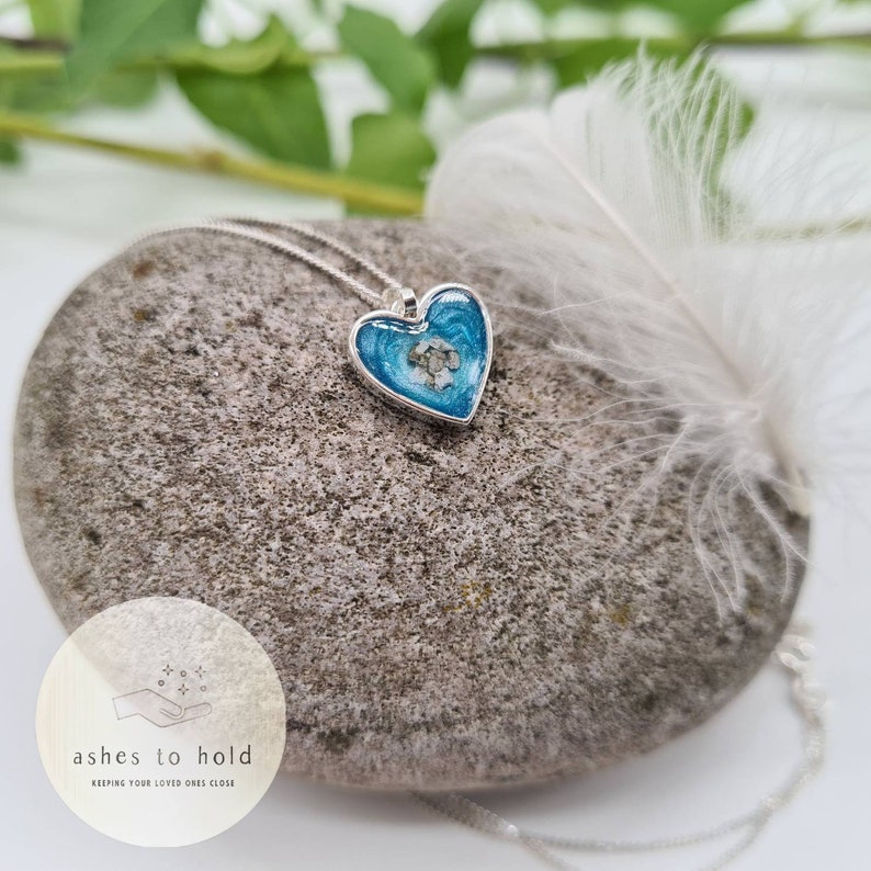 Pode incluir: Um pingente prateado em forma de cora&ccedil;&atilde;o com um centro de resina azul, contendo uma pequena quantidade de material de cor clara. O pingente pende de uma delicada corrente de prata. O colar repousa sobre uma pedra cinza, com uma pena branca por perto.