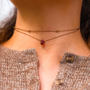 May include: A delicate, layered choker necklace. The top layer features small gold beads, while the lower layer showcases a teardrop-shaped, deep red gemstone pendant. The necklace is worn against a backdrop of a brown knit sweater.