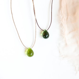May include: Two necklaces with green glass teardrop pendants on a white surface. One necklace has a dark green pendant and the other has a light green pendant.