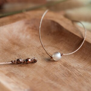 May include: Two necklaces on a wooden surface. One necklace has a single white pearl pendant, the other has multiple small brown beads.