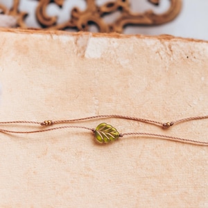 May include: A delicate brown cord necklace with a green leaf charm. The necklace is laying on a vintage book with a worn, cream-colored cover.