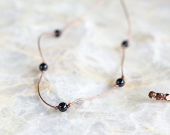 May include: Delicate necklace featuring small, round, black beads spaced along a thin, light brown cord. The necklace has a simple design with a clasp closure. The jewelry is displayed on a light-colored, textured surface.