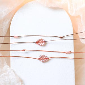 Puede incluir: Primer plano de pulseras con colgantes de hojas. Cada pulsera presenta un pequeño colgante de hoja translúcida de color rosa rojizo en un fino cordón marrón. Las pulseras se exhiben sobre un fondo blanco con una tela suave de color melocotón.