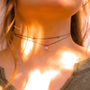 May include: A close-up of a person wearing a layered choker necklace with a small purple gemstone pendant. The necklace is made of thin black cord and the pendant is a light purple colour.