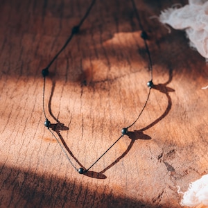 May include: A delicate black necklace with small, dark beads evenly spaced along the chain. The necklace is displayed on a textured, wooden surface, with soft shadows and a white fabric in the background. The necklace is a minimalist jewelry piece.