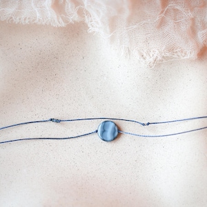 May include: A delicate necklace featuring a small, round, blue-grey stone pendant. The pendant is centered on two strands of fine, navy blue cord. The background is a soft, neutral tone with a light pink fabric draped at the top.