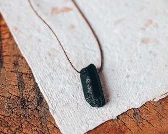 May include: A black crystal pendant necklace with a brown cord. The crystal is rectangular with a rough texture. The necklace rests on a piece of textured, off-white paper, set against a wooden surface.