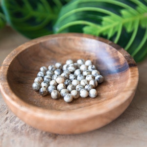 May include: A wooden bowl filled with small, round, speckled beads. The beads are primarily white with gray and brown markings. The bowl is made of light brown wood and sits on a wooden surface, with green leaves in the background.