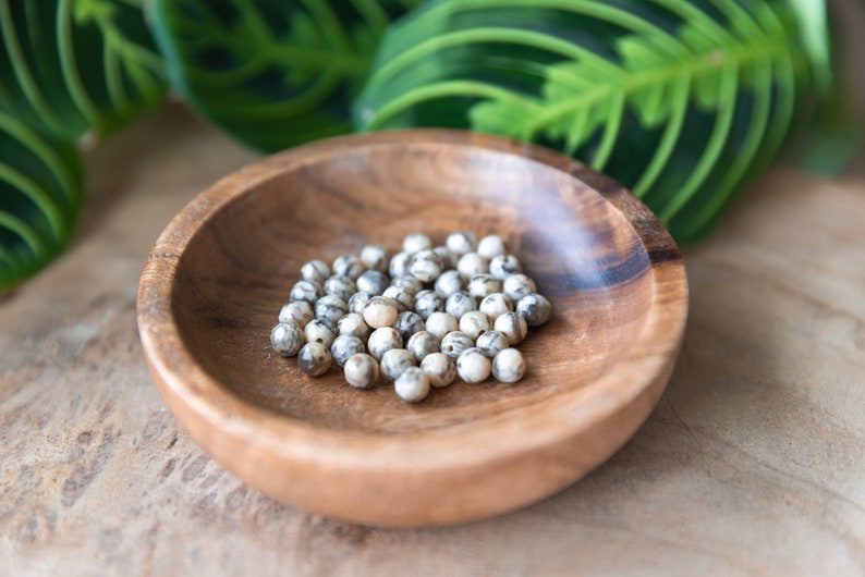 May include: A wooden bowl filled with small, round, speckled beads. The beads are primarily white with gray and brown markings. The bowl is made of light brown wood and sits on a wooden surface, with green leaves in the background.