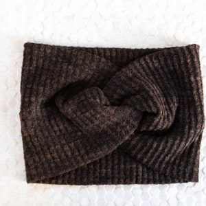 May include: A dark brown, textured headband with a twisted knot design. The headband is made of a soft, woven material and is displayed on a white surface. This accessory is suitable for various hairstyles and adds a touch of warmth.