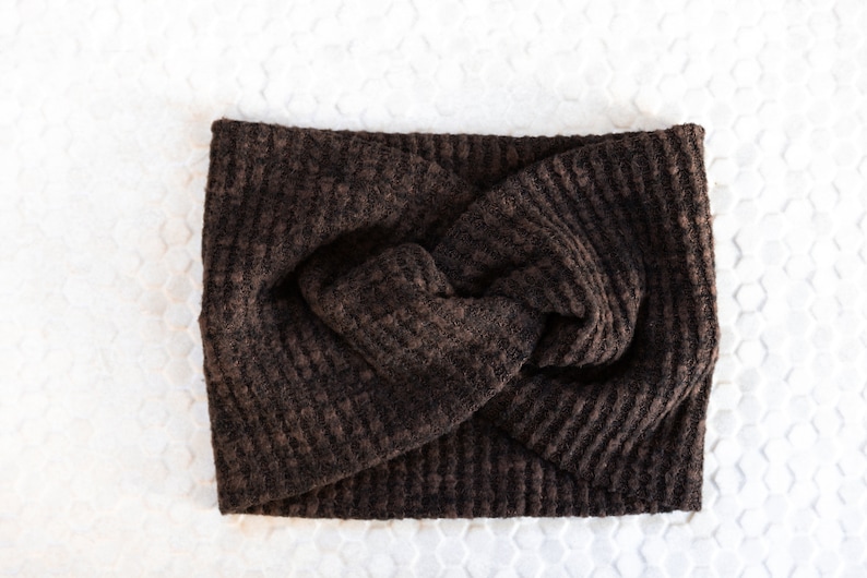May include: A dark brown, textured headband with a twisted knot design. The headband is made of a soft, woven material and is displayed on a white surface. This accessory is suitable for various hairstyles and adds a touch of warmth.