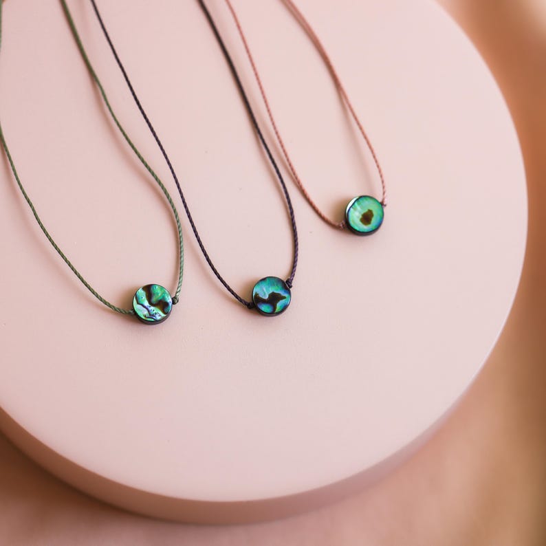 May include: Three delicate necklaces with iridescent abalone shell pendants. Each pendant is a small, round disc with shades of green, blue, and purple. The necklaces have thin, colored cords in green, navy, and brown.