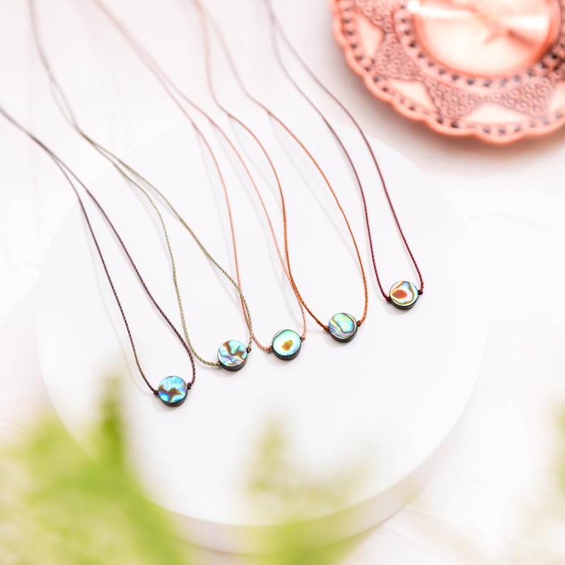 May include: Six delicate necklaces with iridescent abalone shell pendants. Each pendant is round and hangs from a thin, colored cord. The cords vary in color, including brown, green, and purple. The necklaces are displayed on a white surface.