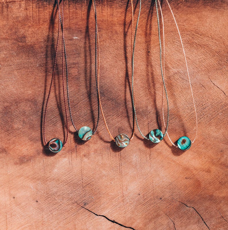 May include: Five necklaces with round, iridescent pendants in shades of teal, brown, and gold. The pendants are strung on thin, brown cords. The necklaces are displayed on a textured, wooden surface.