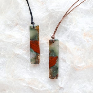May include: Two rectangular pendant necklaces. Each pendant is a mix of green, red, and tan colors. One necklace has a black cord, and the other has a brown cord. The pendants are displayed on a white, textured surface.