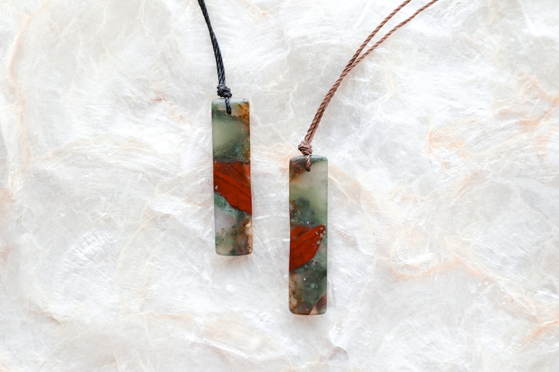 May include: Two rectangular pendant necklaces. Each pendant is a mix of green, red, and tan colors. One necklace has a black cord, and the other has a brown cord. The pendants are displayed on a white, textured surface.