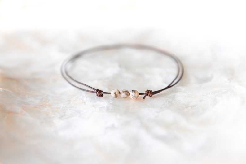 May include: A minimalist bracelet featuring three speckled beads strung on a thin, dark brown cord. The cord is knotted at each end, creating a simple, elegant design. The bracelet is shown on a white, textured surface.