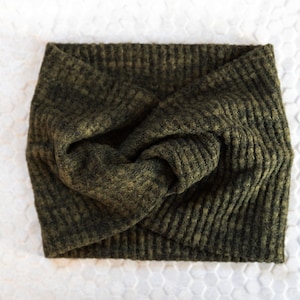 May include: Olive green knitted headband with a twisted knot design. The headband is made of a textured, waffle-knit fabric. The accessory is shown on a white surface with a honeycomb pattern.