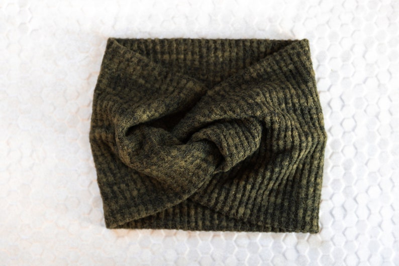 May include: Olive green knitted headband with a twisted knot design. The headband is made of a textured, waffle-knit fabric. The accessory is shown on a white surface with a honeycomb pattern.