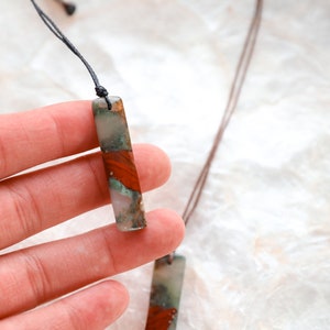 May include: A rectangular pendant necklace with a natural stone appearance. The pendant features a mix of red, green, and white colors. The necklace is strung on a dark brown cord. The background is a light, textured surface.