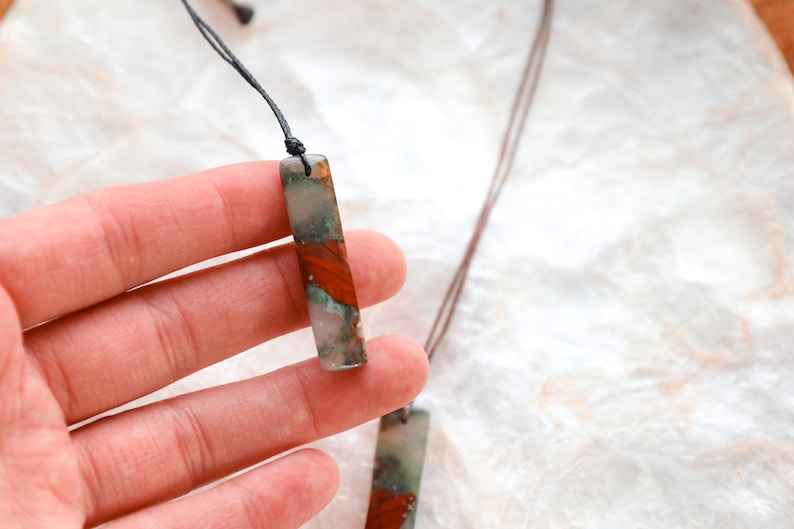 May include: A rectangular pendant necklace with a natural stone appearance. The pendant features a mix of red, green, and white colors. The necklace is strung on a dark brown cord. The background is a light, textured surface.