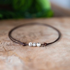 May include: A minimalist bracelet featuring three speckled, off-white beads strung on a thin, dark brown leather cord. The cord is knotted at each end. The bracelet is displayed on a wooden surface.